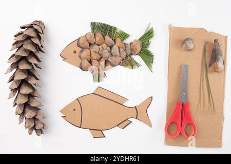 Fai da te, pesca artigianale con scaglie di pinecone, aghi di pino e cartone con forbici rosse su sfondo bianco. Composizione piatta per la lavorazione, riciclo Foto Stock