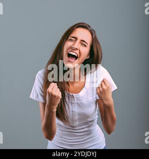 Giovane donna entusiasta con lunghi capelli castani. Indossa una maglietta bianca. Esprime la sua eccitazione urlando e alzando i pugni contro una schiena grigia Foto Stock