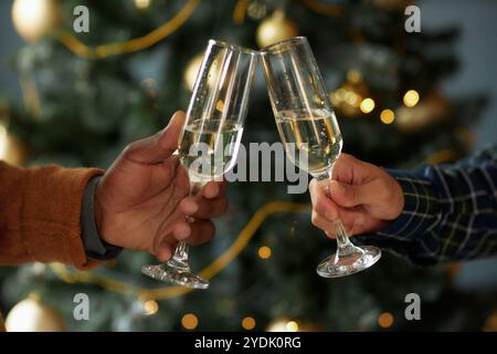 Vista laterale di due amici maschi che brindono bicchieri di champagne per festeggiare le vacanze mentre sorseggiano un drink e trascorrono il tempo insieme la vigilia di Natale con le luci della ghirlanda sullo sfondo, copia spazio Foto Stock