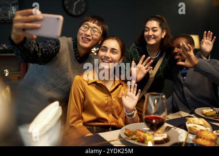 Foto sincera di un uomo asiatico che fa selfie con quattro allegri amici che sventolano le mani alla fotocamera Foto Stock