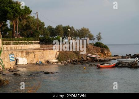 Casa su una spiaggia rialzata con piccole barche ormeggiate e tre uscite dall'acqua, vicino a una bandiera greca dipinta su un muro di cemento. L'erba è secca. Foto Stock