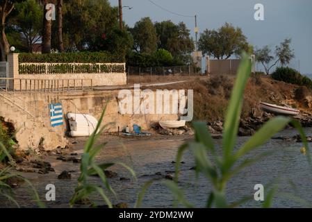 Casa su una spiaggia rialzata con piccole barche ormeggiate e trascinate fuori dall'acqua, vicino a una bandiera greca dipinta su un muro di cemento. Piante in primo piano. Foto Stock