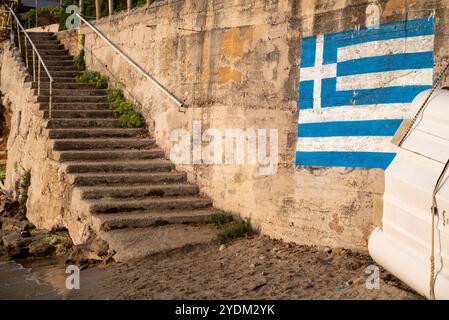Bandiera greca dipinta su un muro di cemento con scalinata in pietra e ringhiere metalliche che si dirigono dalla spiaggia sabbiosa a una casa. Una barca è stata trascinata fuori dall'acqua. Foto Stock