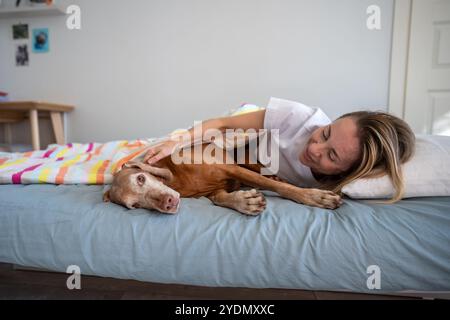 Ridendo donna rilassata che giace con il cane Magyar Vizsla a letto, pigra, non vuole andare da nessuna parte la mattina Foto Stock