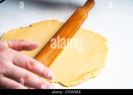 Le mani degli uomini stenderanno l'impasto corto con un mattarello su un tavolo bianco. Il fornaio sta lavorando con l'impasto. Ricetta per preparare una torta o dei biscotti. Ciao Foto Stock