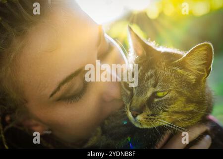 Retroilluminata dalla luce del sole dorata, una giovane donna bacia dolcemente il suo gatto tabby, creando una scena commovente di amore e connessione tra uomo e animale Foto Stock