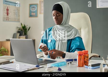 Medico afroamericano musulmano seduto alla scrivania, che prescrive farmaci mentre tiene in mano una bottiglia, con cartelle mediche sulle pareti, focalizzato sullo schermo del computer portatile Foto Stock