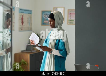 Indossando un velo, una donna musulmana afroamericana che studia attentamente i documenti in un ufficio moderno e ben illuminato con carte mediche alle pareti Foto Stock