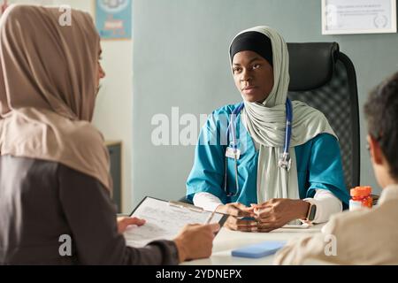 Fornitore di assistenza sanitaria musulmana che interagisce con i pazienti, discutendo diagnosi medica, indossando uno stetoscopio e scrub medici, seduto in un ufficio clinico, focalizzato sulla cura del paziente Foto Stock