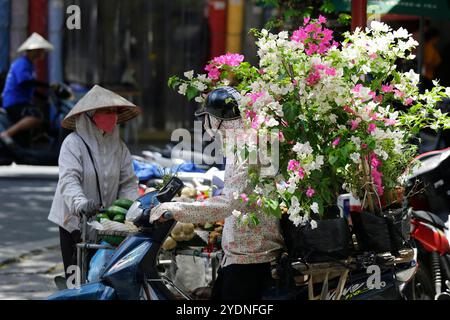 Hanoi, Vietnam - 23 luglio 2023: Il venditore di fiori vietnamita guida la sua moto attraverso una strada trafficata. Foto Stock