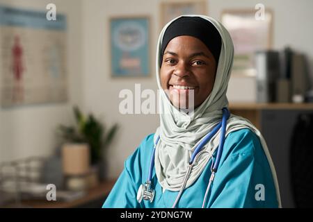 Medico donna musulmano sorridente in abbigliamento medico in piedi in clinica e guardare contenuto. Lo sfondo mostra poster e attrezzature mediche che aggiungono un'atmosfera professionale Foto Stock