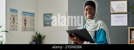 Giovane donna musulmana afroamericana in piedi in ambiente sanitario sorridente calorosamente mentre tiene gli appunti e indossa un abbigliamento professionale con certificati sul muro in background Foto Stock