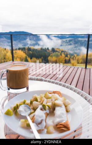 Pancake con frutta tritata e tazza di caffè sullo sfondo mozzafiato del paesaggio autunnale. Colazione in montagna. Colazione su una terrazza in legno in montagna Foto Stock