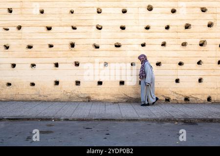 Composizione di una donna anziana che cammina lungo la strada vestita di una djellaba grigia di fronte alle massicce mura cittadine di Fez in Marocco, Nord Africa Foto Stock