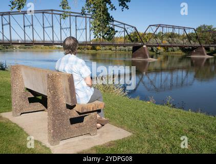 Donna anziana seduta su una panchina ammirando lo storico Fort Benton Bridge che attraversa il fiume Missouri nel Montana. Foto Stock