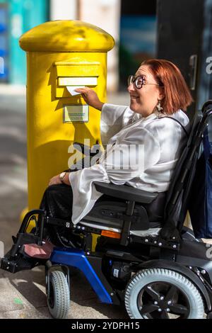 Donna in sedia a rotelle che pubblica una lettera nella casella postale pubblica gialla Foto Stock