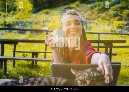 La giovane donna freelance sorridente con dreadlock, che indossa una camicia a quadri rossa e nera, accarezza il suo gatto mentre beve caffè e lavora sul suo lapto Foto Stock