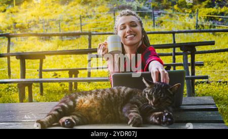 Freelance sorridente con dreadlock, camicia rossa, caffè, gatto degli animali domestici, lavora su un computer portatile all'aperto, godendosi la natura e la compagnia degli animali Foto Stock