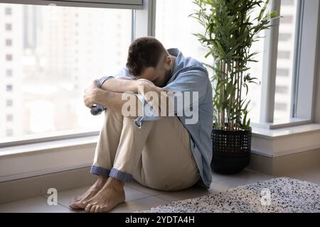 Depresso giovane uomo infelice seduto alla finestra sul pavimento Foto Stock