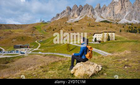 Un escursionista solitario fa una pausa su un arroccato per ammirare i panorami mozzafiato delle Dolomiti, circondati da un vivace fogliame autunnale e da vette maestose che raggiungono le nuvole. Passo Rolle, Trento Foto Stock