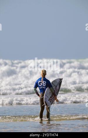 Una surfista con una maglietta GB Surfing sta in acqua, tenendo la tavola, contemplando le onde prima di uscire per fare surf. Foto Stock