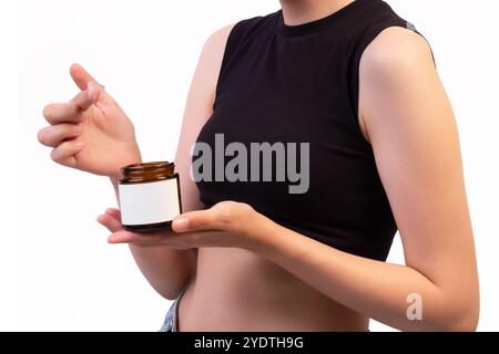 Donna che tiene in mano un vaso di crema aperto in un contenitore color ambra con etichetta vuota – presentazione dei prodotti per la cura della pelle e la bellezza per il branding e la pubblicità Foto Stock