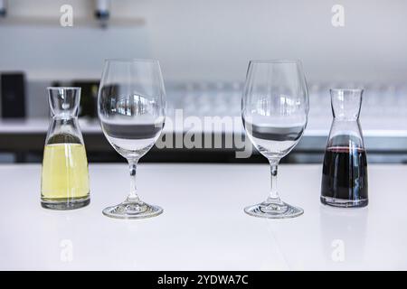 Due bicchieri da vino vuoti e un paio di piccoli decanter ripieni di vini bianchi e rossi, assaggia calici su un luminoso bancone bianco della sala di degustazione Foto Stock