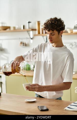 Un bel giovane con capelli ricci versa il caffè appena preparato in una tazza, godendosi un momento nella sua cucina. Foto Stock