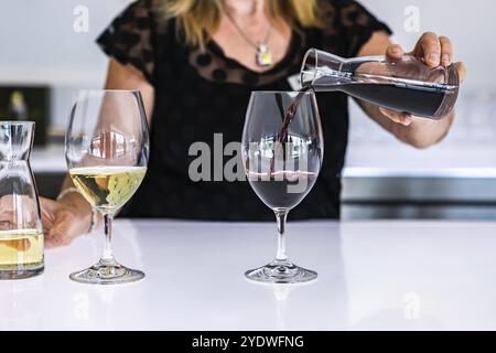 La barista donna si avvicina mentre versa vino rosso da un piccolo decanter al wineglass, accanto a un bicchiere di vino bianco, luminoso bancone da bar Foto Stock