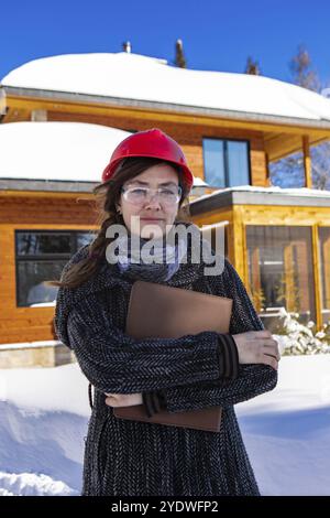 Ritratto verticale di un architetto edile donna che indossa un elmetto rosso e tiene in mano gli appunti. lei è in piedi sul fronte di un edificio coperto di neve Foto Stock