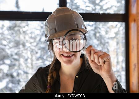 Lavoratrice caucasica che sorride. ispettore edile che guarda attraverso una lente d'ingrandimento. indossava un elmetto grigio durante l'ispezione della qualità dell'aria domestica Foto Stock