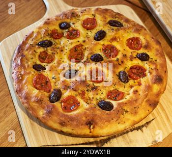 Pane piatto appena sfornato con pomodori e olive, posto su un asse di legno bruciato. Deliziosa focaccia spessa con crosta croccante. Pizza rotonda alta Foto Stock