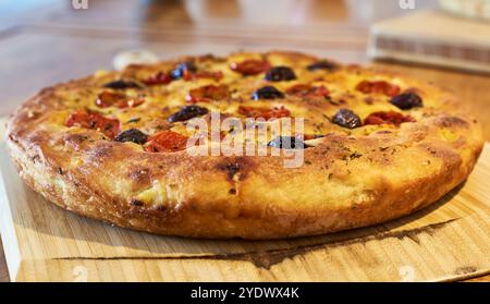Primo piano di focaccia pugliese al forno con una crosta marrone dorata, ricoperta di fette di pomodoro, olive nere ed erbe aromatiche. Deliziosa pizza cotta al forno Foto Stock