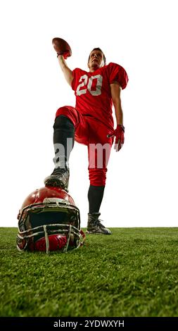 Campione. Vista dal basso dell'uomo, giocatore di football americano in uniforme rossa, in piedi con la palla, posizionamento della gamba sul casco disteso sullo stadio. Foto Stock