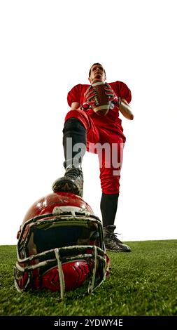 Campione. Vista dal basso dell'uomo, giocatore di football americano in uniforme rossa, in piedi con la palla, posizionamento della gamba sul casco disteso sullo stadio. Foto Stock