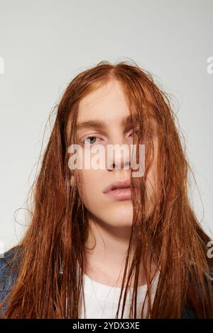 Un ragazzo adolescente mostra i suoi lunghi capelli rossi e l'abito in denim mentre posa con sicurezza in uno studio. Foto Stock