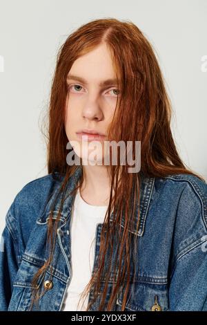 Un ragazzo adolescente sfoggia i suoi lunghi capelli rossi e l'abito in denim, esprimendo il suo stile unico in studio. Foto Stock