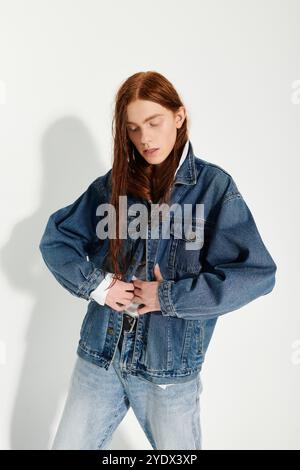 Un ragazzo adolescente sfoggia i suoi lunghi capelli rossi e l'abito in denim alla moda mentre posa con sicurezza in uno studio. Foto Stock