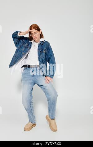 Un ragazzo adolescente sfoggia i suoi lunghi capelli rossi e l'abbigliamento in denim, creando una posa alla moda in uno studio. Foto Stock