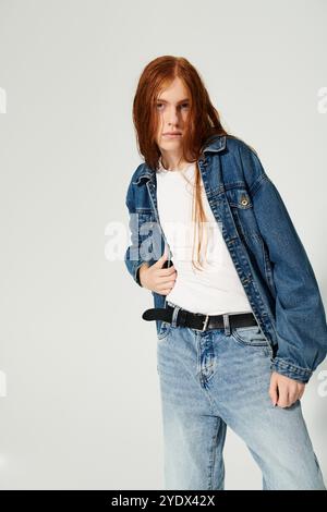 Un adolescente con lunghi capelli rossi si staglia con sicurezza in un outfit denim, che irradia creatività e stile. Foto Stock