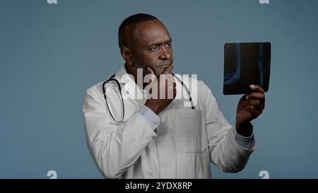 Uomo afroamericano pensivo e premuroso medico radiologo maschio etnico chirurgo in studio sfondo blu guardare radiografia risultato controllo Foto Stock