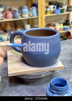 Il processo di pittura di un vaso di argilla durante una masterclass di ceramica. Artigianato e creatività in azione. Il vaso è ricoperto di smalto blu, creato Foto Stock