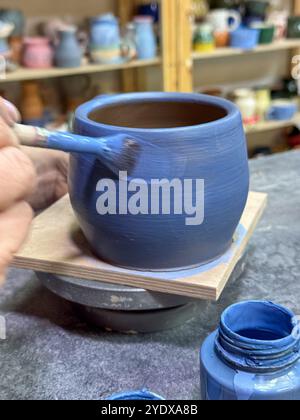 Il processo di pittura di un vaso di argilla durante una masterclass di ceramica. Artigianato e creatività in azione. Il vaso è ricoperto di smalto blu, creato Foto Stock