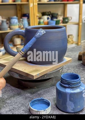 Il processo di pittura di un vaso di argilla durante una masterclass di ceramica. Artigianato e creatività in azione. Il vaso è ricoperto di smalto blu, creato Foto Stock