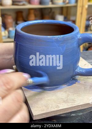 Il processo di pittura di un vaso di argilla durante una masterclass di ceramica. Artigianato e creatività in azione. Il vaso è ricoperto di smalto blu, creato Foto Stock