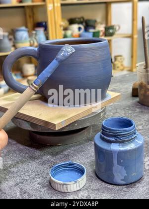 Il processo di pittura di un vaso di argilla durante una masterclass di ceramica. Artigianato e creatività in azione. Il vaso è ricoperto di smalto blu, creato Foto Stock