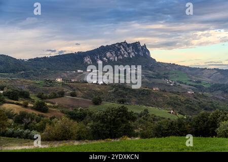 Vista panoramica del paesaggio collinare con l'impressionante massiccio roccioso delle tre torri iconiche di San Marino sullo sfondo. Le ripide pendici della montagna sono intervallate da terrazze boscose. Piccoli villaggi e case individuali sono sparsi sulle pendici. Il paesaggio è un misto di campi coltivati e foreste in varie sfumature di verde. Via del progresso, Montegiardino, San Marino Foto Stock