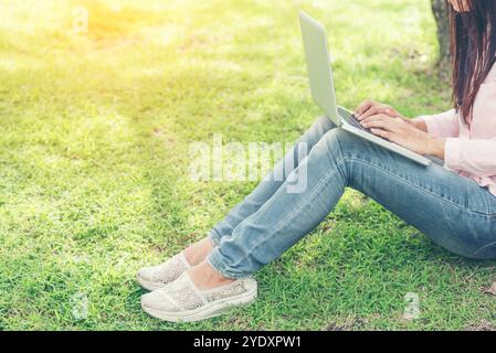 Donna asiatica seduta parco verde con computer portatile. Donna che lavora in un'azienda felice di un notebook che utilizza un notebook con le mani che digitano sulla tastiera Foto Stock