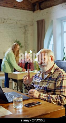 Uomo anziano, professionista, seduto in uno spazio di lavoro collaborativo, alla scrivania con notebook che ricerca, esamina report o analisi. Foto Stock