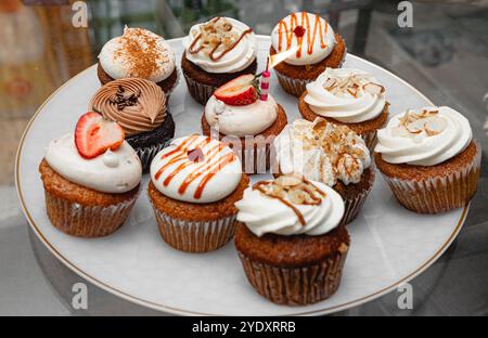 Un piatto decorativo con un assortimento di cupcake ricoperti di glassa, fette di frutta e gocciolatoi, che mostra vari gusti e disegni per un dedante Foto Stock